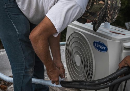 A technician performs maintenance on an outdoor air conditioning unit, focusing on hose connections.